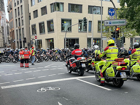 Giro 2025 Rettungskräfte auf Motorrädern stehen in Position, auf der Straße ist ein Feld von Radfahrer*innen. Zwei Einsatzkräfte koordinieren die Situation. Im Hintergrund ist ein Kirchturm zu sehen.