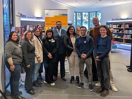 Gruppenfoto Eine Gruppe von Menschen steht vor einem "Wochen gegen Rassismus" Banner, im Hintergrund sind Bücherregale zu sehen.