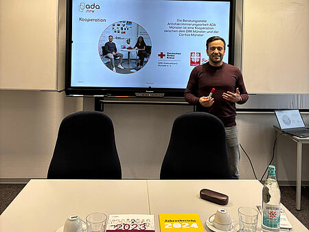 ADA Workshop Ein Mann steht vor einer Präsentation und hinter einem Tisch, auf dem Informationsmaterialen liegen.