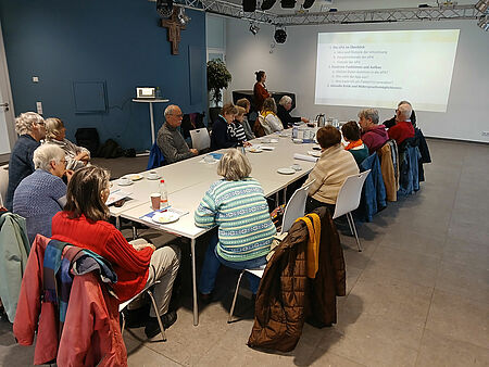 Fit für den digitalen Alltag Eine Gruppe von älteren Menschen sitzt um einen Tisch herum und schaut auf eine Präsentation an der Wand, neben der eine Referentin steht.