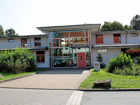Wolkenburg Ein modernes, zweistöckiges Gebäude mit großen Fenstern und einer roten Eingangstür. Ein gepflasterter Weg führt durch grüne Büsche und Bäume zum Eingang.