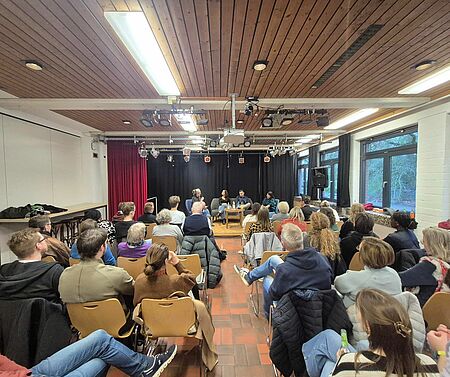 Publikum In einem großen Raum sitzen viele Menschen in Sitzreihen. Vor ihnen sitzen mehrere Personen auf einem Podium.