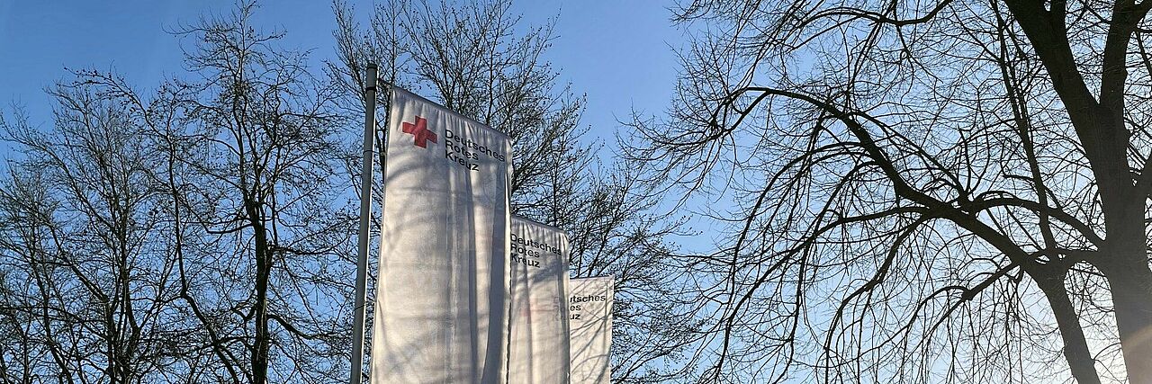 Fahnen Winter Drei weiße Fahnen mit DRK-Logo vor einem Baum und blauem Himmel.