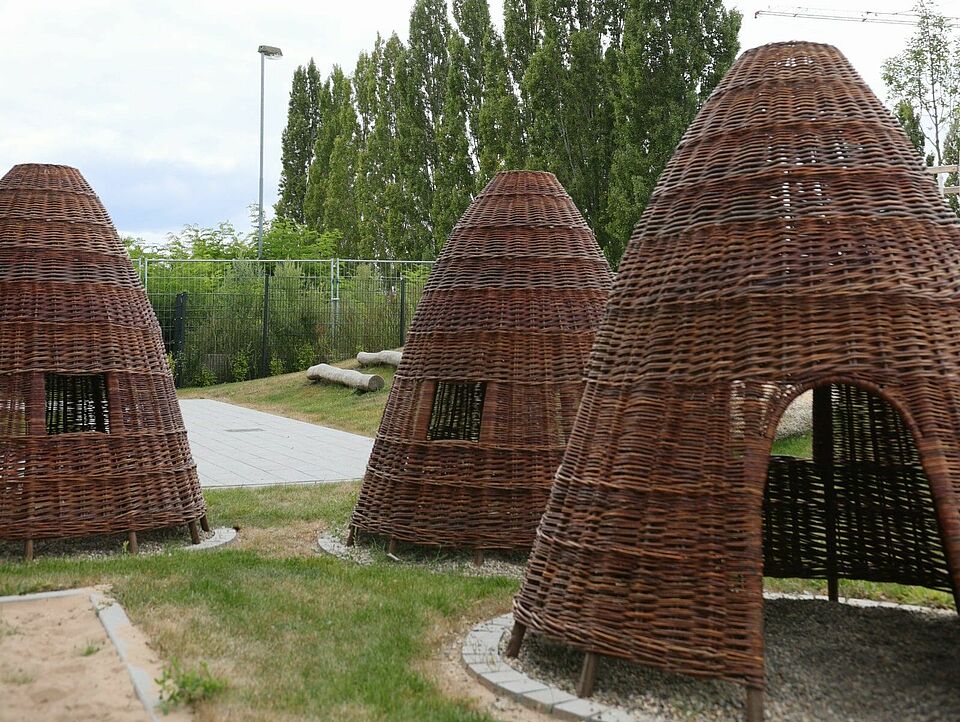 Hütten Mehrere kegelartige Hütten aus geflochtenen Weidenruten auf einer Wiese, umgeben von Bäumen und einem Zaun unter leicht bewölktem Himmel.