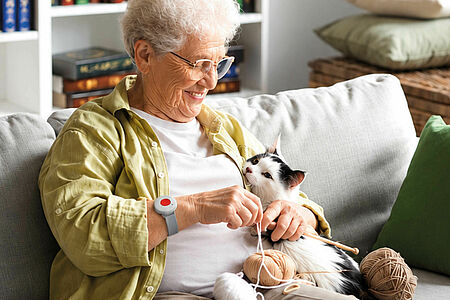 Hausnotrufwochen Stricken Eine Seniorin sitzt mit einer Katze auf dem Sofa und strickt. Dabei trägt sie einen roten Hausnotruf-Sender am Handgelenk.