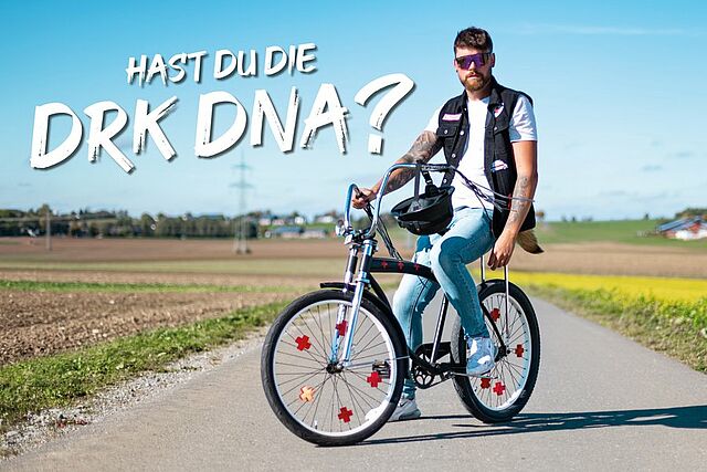 Biker Eine Person fährt mit dem Fahrrad auf einer Landstraße umgeben von Feldern und einigen Gebäuden im Hintergrund. In den Speichen befinden sich Rotkreuzreflektoren. Text im Bild: „Hast du die DRK DNA?“