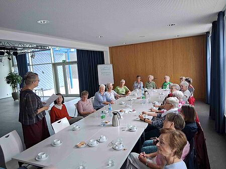 Mental und Gedächtnistraining Eine Gruppe von älteren Leuten sitzt um einen großen Tisch, auf dem Getränke und Geschirr stehen, herum. Im Vordergrund steht eine Fraz und spricht zur Gruppe.