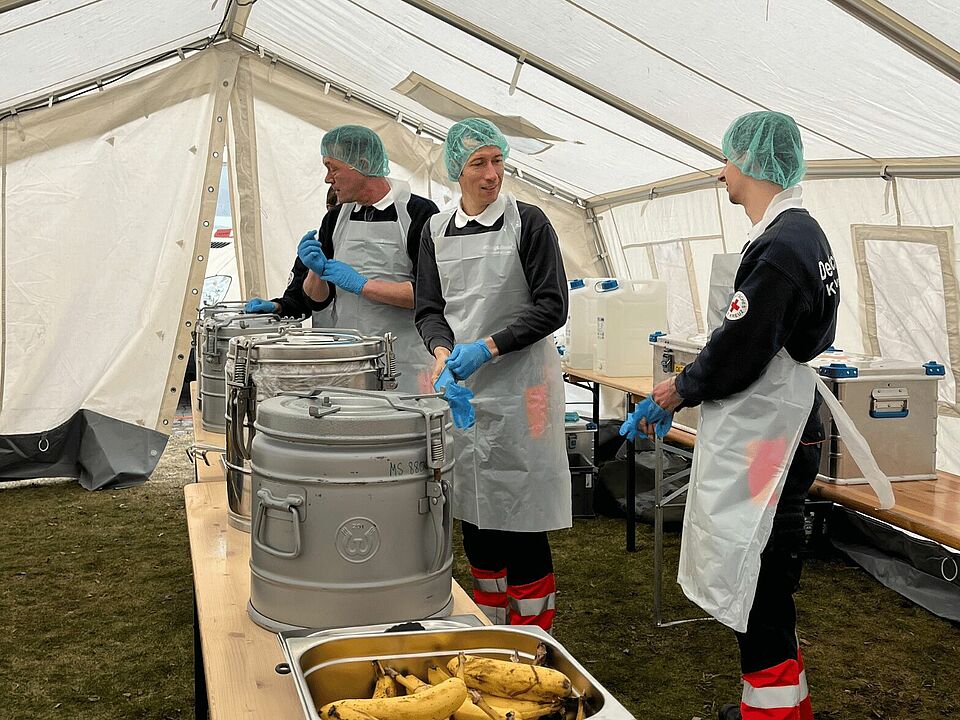 Drei Ehrenamtliche in Schutzkleidung stehen vor einem langen Tisch mit Essen