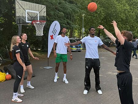 Fit in Berg Fidel Fünf junge Menschen stehen vor einem Basketballkorb und werfen einen Basketball. Im Hintergrund ist eine DRK-Beachflag zu sehen.