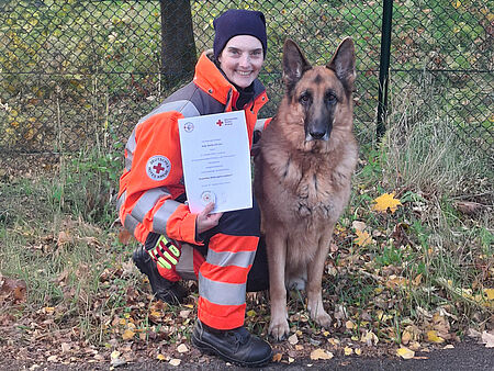 Anja und Leo Eine DRK-Einsatzkraft kniet neben einem Hund mit einer Urkunde in der Hand.