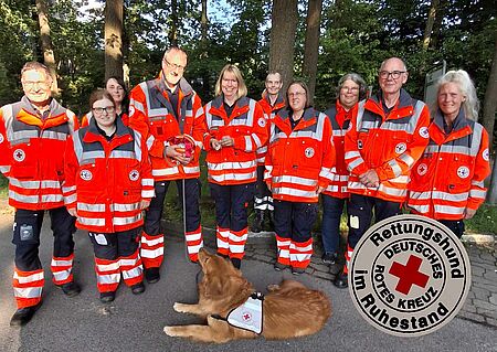 Lupus Eine Gruppe von Personen in DRK-Einsatzkleidung, im Vordergrund liegt ein Hund. Rechts auf dem Bild ist eine Darstellung der DRK-Rettungshund im Ruhestand Plakette zu sehen