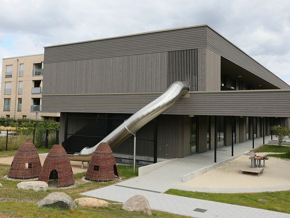 Außengelände Modernes Gebäude mit Außengelände. Eine Rutsche führt vom ersten Stock runter in den Garten. Zu sehen ist ein Sandbereich, drei kleine Hütten aus Naturmaterialien und zahlreiche Steine und Baumstämme.