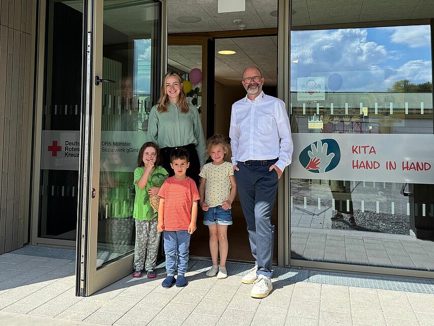 Neueröffnung Kita Hand in Hand Zwei Personen mit drei Kindern stehen in einer Tür von einem neuen Gebäude. Auf den Glasfronten ist das Rotkreuzlogo und das Logo der Kita Hand in Hand zu sehen.