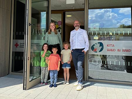 Neueröffnung Kita Hand in Hand Zwei Personen mit drei Kindern stehen in einer Tür von einem neuen Gebäude. Auf den Glasfronten ist das Rotkreuzlogo und das Logo der Kita Hand in Hand zu sehen.